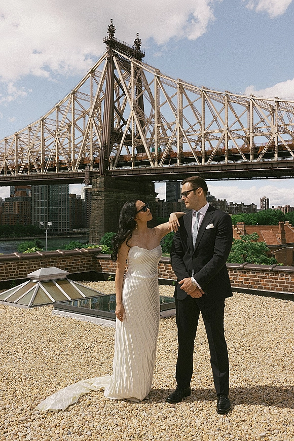 Couple portrait of bride in a strapless wedding dress and groom in black suit with sunglasses on a rooftop by a bridge and city skyline