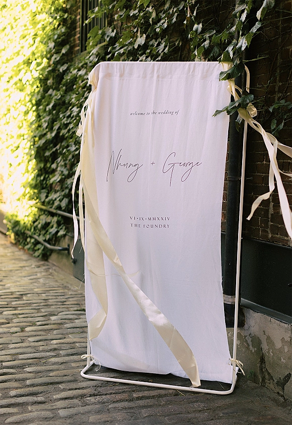Wedding welcome sign in fabric with ribbon ties on a metal stand, set against an ivy and brick wall along a cobblestone alleyway