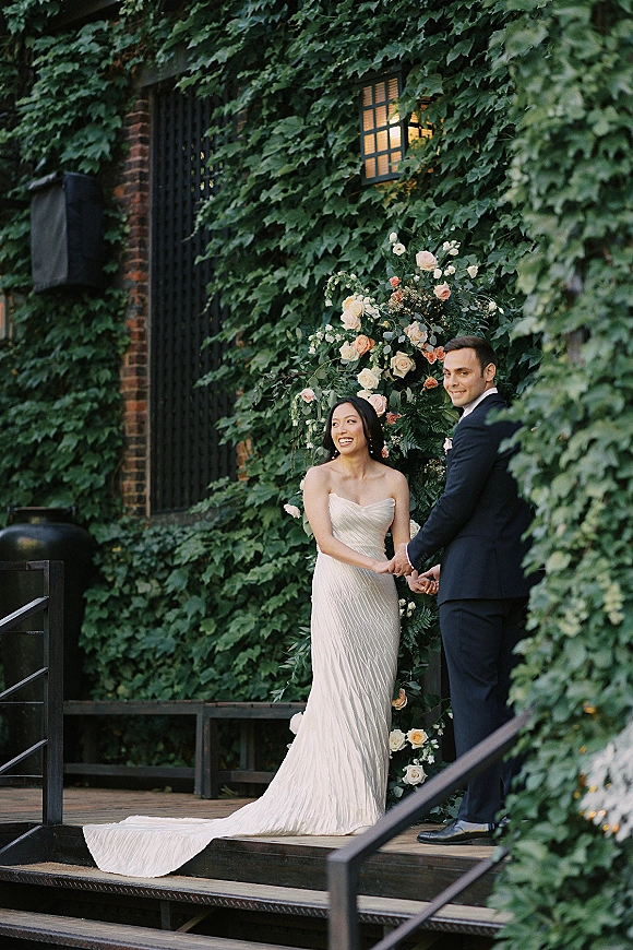 Couple portrait of bride and groom holding hands under a floral arch with roses and greenery, posed by an ivy-covered brick wall outdoors
