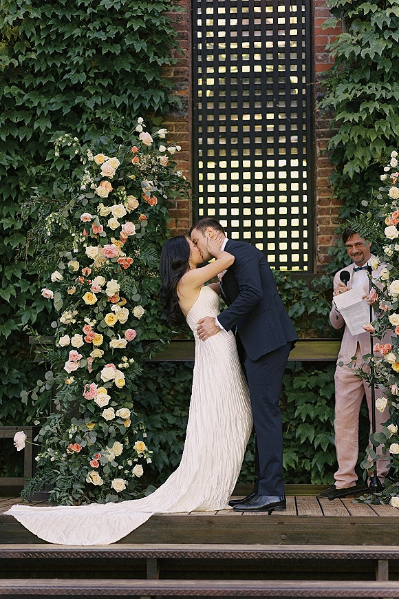 Wedding kiss as bride and groom embrace beneath a rose and greenery arch, with officiant and ceremony papers against an ivy brick wall