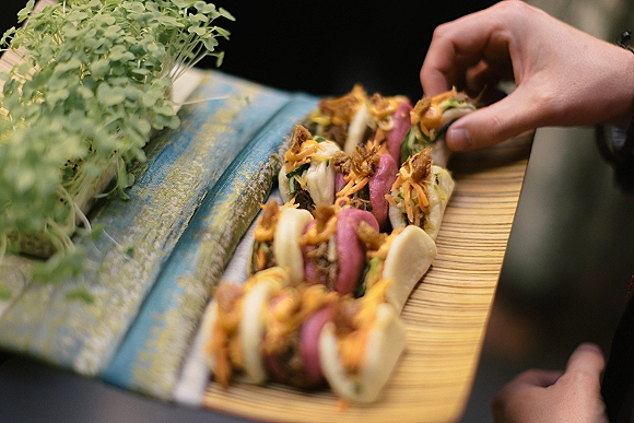 Wedding catering platter of bao buns with greenery accents as hands pass bite-size appetizers over a fabric runner at cocktail hour