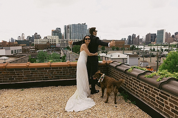 Couple portrait in sunglasses, bride hugging groom in a strapless wedding dress as he opens his arms on a rooftop terrace with city skyline