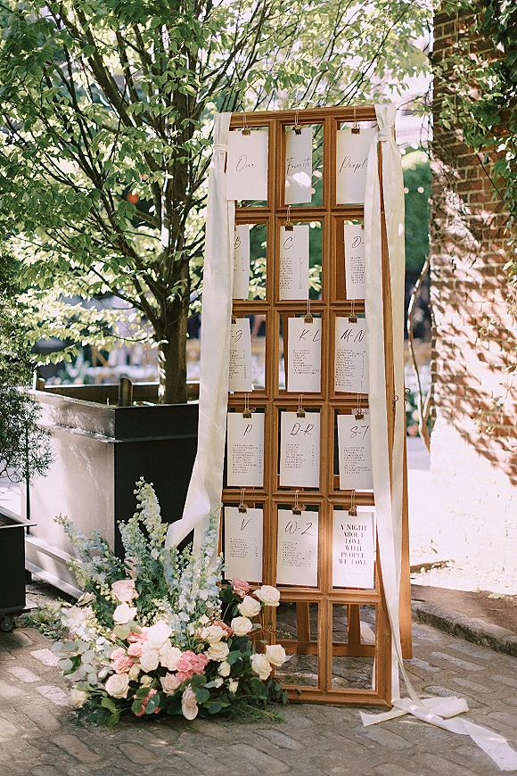 Wedding seating chart on a wood window frame with calligraphy escort cards, ribbon draping, and rose greenery accents on a brick walkway
