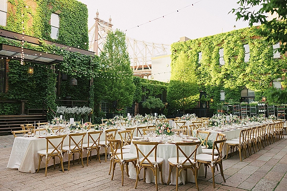 Outdoor reception decor with long banquet and round tables in white linens, cross back chairs, floral centerpieces, and string lights in an ivy-brick courtyard