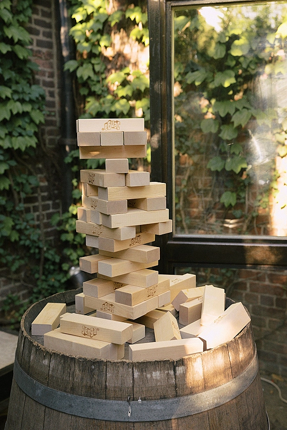 Giant jenga game set for wedding lawn games on a patio, stacked wooden blocks beside a wooden barrel table by ivy brick wall