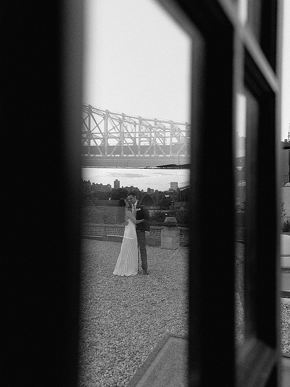 Couple portrait of bride and groom embracing, her wedding dress and his suit with boutonniere on a rooftop terrace with city skyline and bridge behind