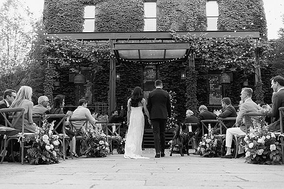 Processional with bride walking down aisle beside groom, aisle florals and lanterns under string lights in an ivy courtyard, black and white