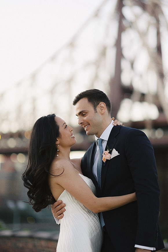 Couple portrait of bride in a strapless dress and groom in a suit embracing on a bridge, city skyline and waterfront bokeh lights behind them