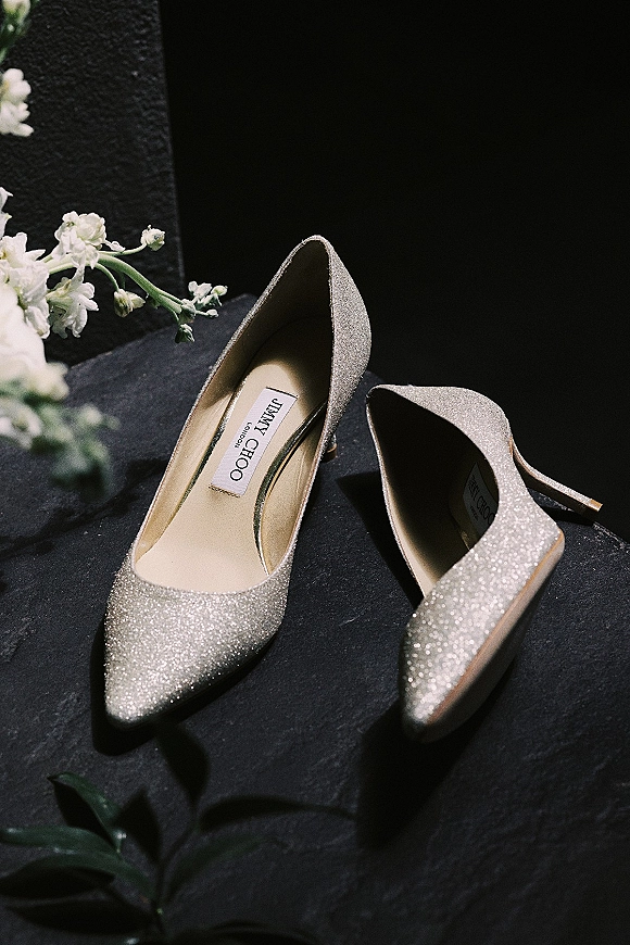 Wedding shoes with glitter wedding heels, pointed-toe sparkle pumps styled with floral stems on a dark fabric flatlay backdrop