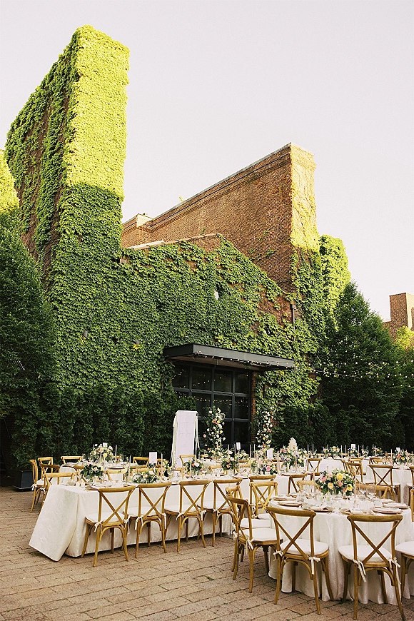 Outdoor reception decor with long banquet tables, white linens, floral centerpieces and candles under bistro lights in an ivy brick courtyard patio