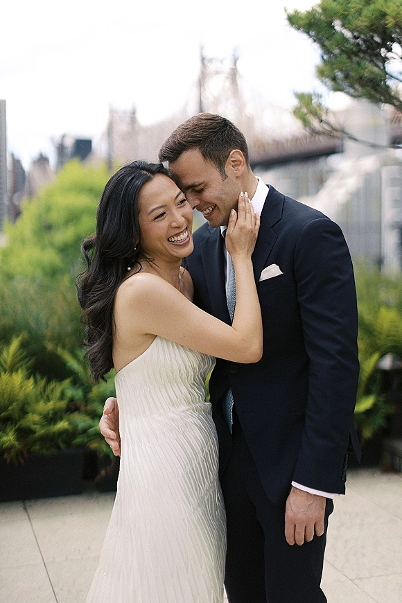 Couple portrait of bride and groom laughing as she cups his face, her strapless gown and his navy suit on a terrace with skyline and bridge behind