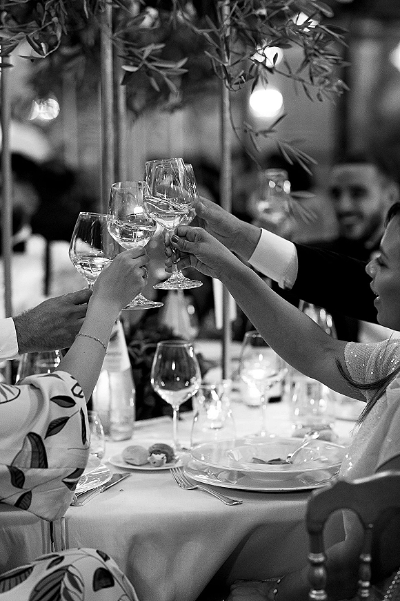 Wedding toast as guests clink champagne flutes over a candlelit dinner table with greenery centerpiece, blurred reception room and bistro lights behind