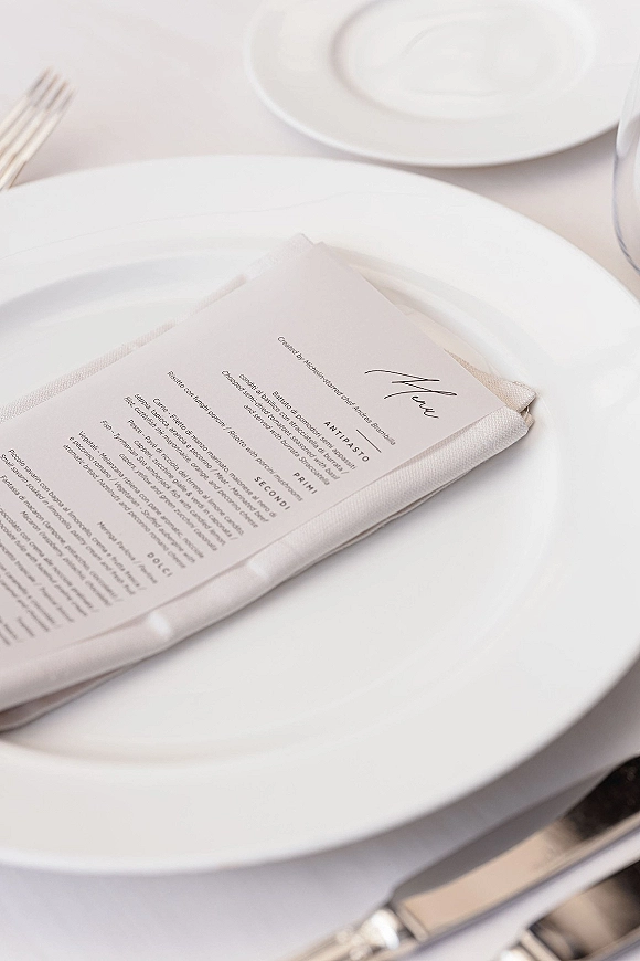 Wedding place setting with a wedding menu card on a folded linen napkin, layered white plates, silver flatware, and wine glass on white tablecloth