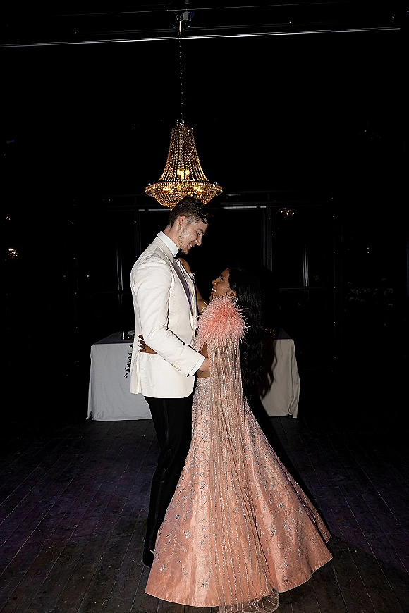 Wedding couple portrait of bride and groom embrace, her blush embellished gown and feather stole under a chandelier in a dark reception room