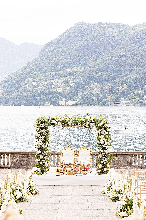Ceremony setup with an outdoor ceremony arch of pink and white flowers on a stone terrace overlooking a lake and mountains, with gold chairs