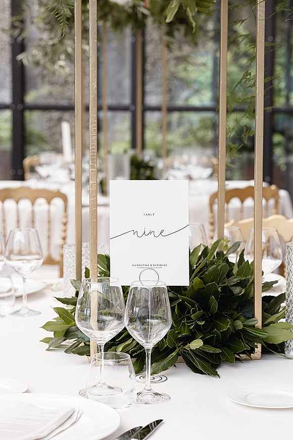 Reception tablescape with wedding table number in a gold frame, greenery garland, white plates, and glassware by large windows overlooking greenery