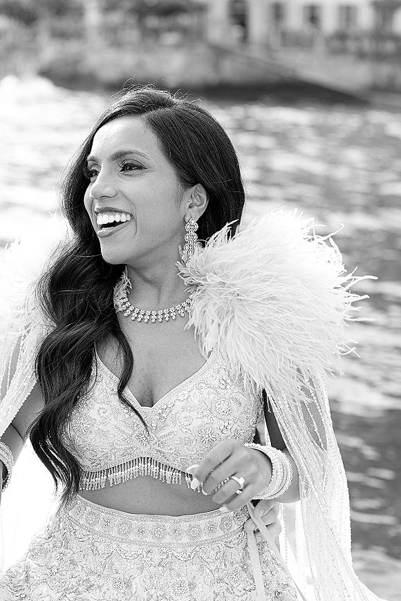 Bridal portrait of a laughing bride in a beaded lehenga with feather stole by river water near a bridge in black and white