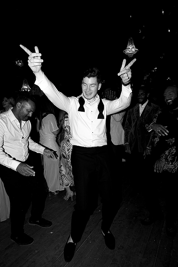 Wedding reception dancing as guests in suits and cocktail dresses throw hands up on a wood dance floor under chandelier lighting in a dark room