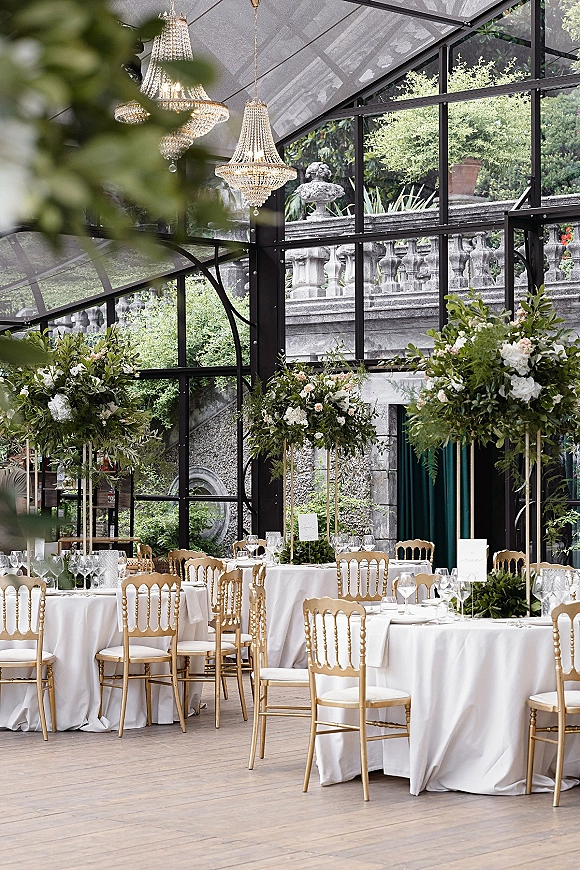 Reception tablescape with round white linens, tall blush-and-white floral centerpieces, and gold Chiavari chairs under crystal chandeliers in a glass greenhouse