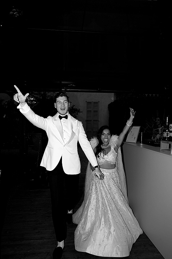 Reception entrance as couple holds hands, groom in white dinner jacket and bow tie beside bride in lehenga on a wooden deck at night