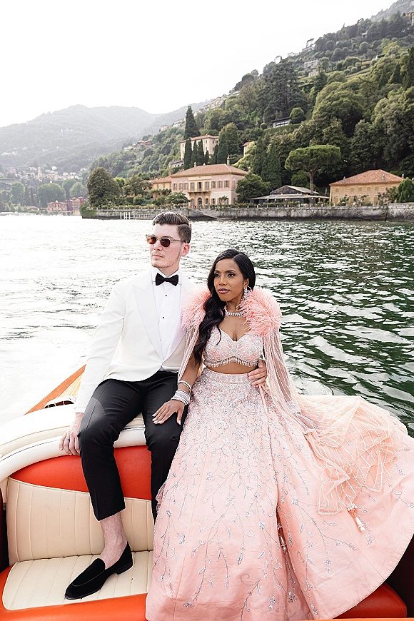 Couple portrait of bride in embroidered lehenga and veil beside groom in white tuxedo and sunglasses on a speedboat with mountains and villas behind