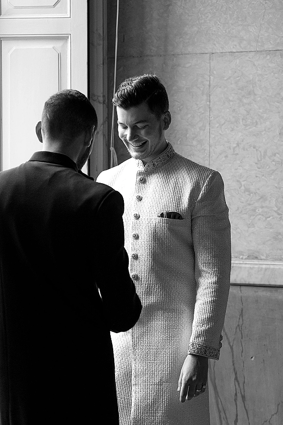 Groom getting ready in an ivory sherwani with embroidered collar and pocket square, adjusting buttons in soft window light by a paneled door