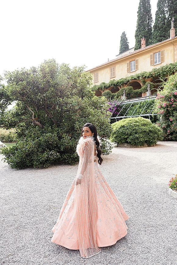 Bridal portrait of a bride in a pink wedding dress with a sheer beaded cape and feather trim, looking over her shoulder in a villa garden courtyard