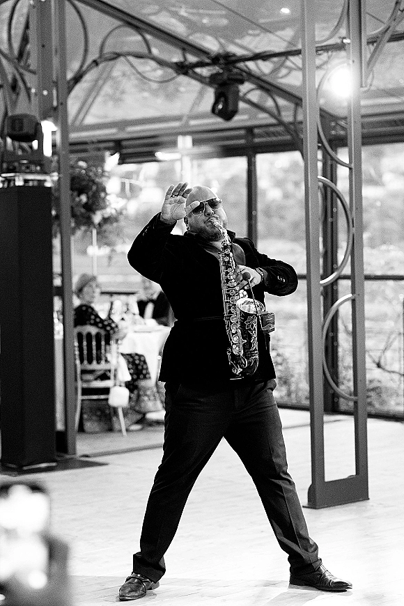 Wedding band performance with a wedding saxophonist in sunglasses playing sax on stage at a glass-walled reception with guests and tables