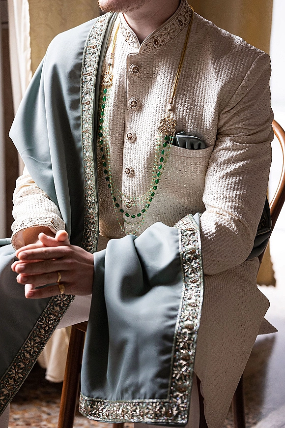 Groom portrait in a sherwani groom outfit, seated by window light, showing embroidered sherwani, shawl drape, beaded necklace and ring