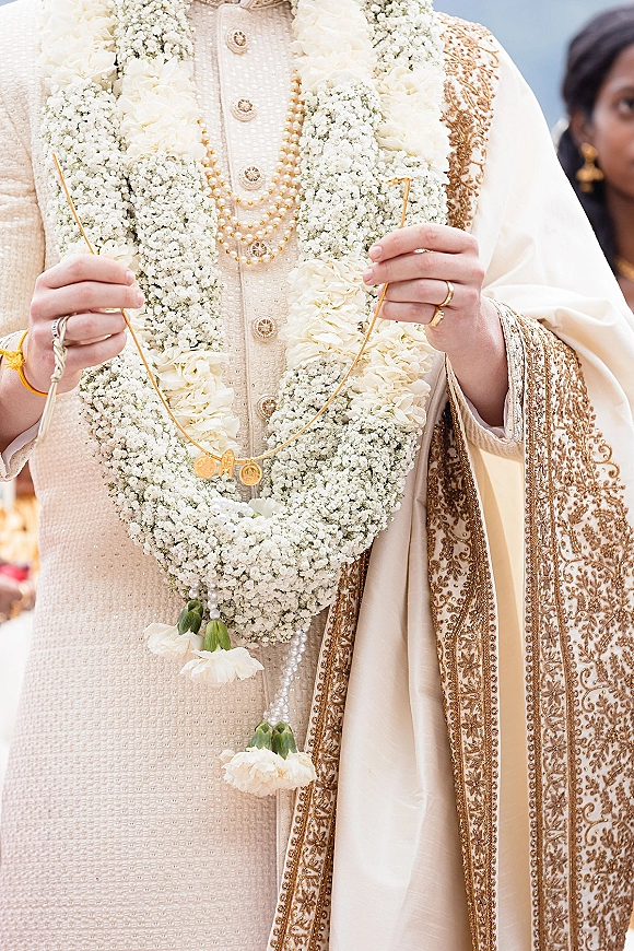 Wedding garland from the jaimala ceremony draped over an embroidered sherwani with pearl and gold necklaces, guests blurred behind