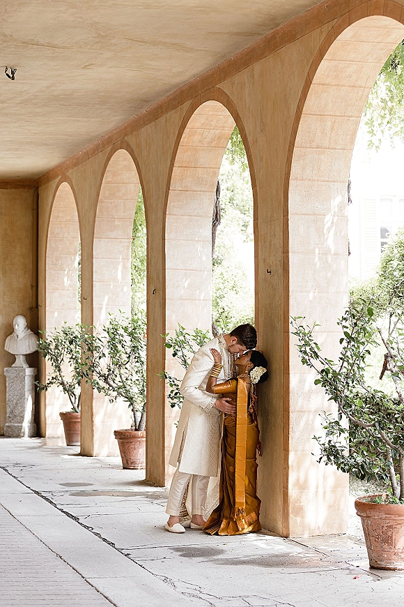 Wedding kiss portrait of an Indian wedding couple embracing, bride in silk sari with floral hair garland under an arched colonnade