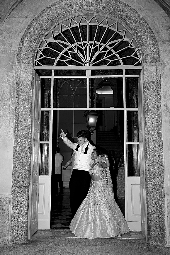 Wedding reception exit as newlyweds leave reception through an arched stone doorway, bride in lehenga laughing beside groom in tuxedo amid guests