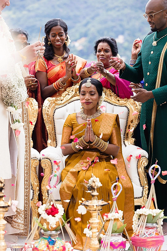 Hindu wedding ceremony as bride in gold sari and groom in sherwani sit on ornate white-gold thrones amid petal shower outdoors with mountains
