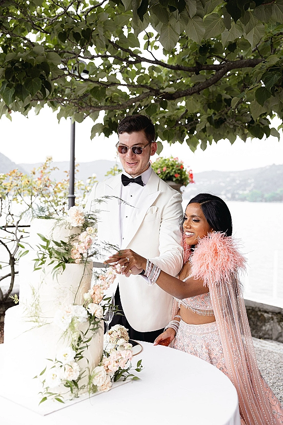 Wedding cake cutting as bride in embellished outfit and groom in white tuxedo slice floral cake by a lake with mountains beyond