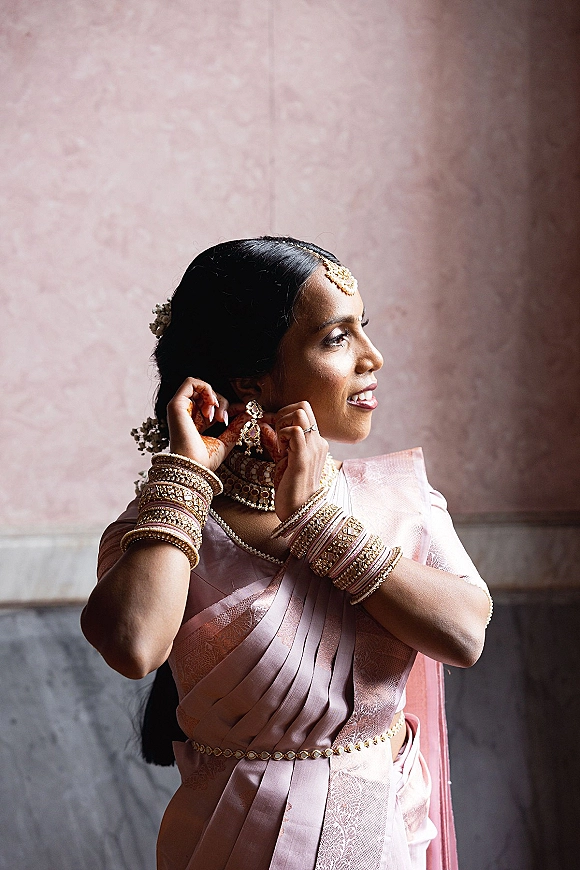 Bride portrait of a South Asian bride in a pink silk saree with gold bangles and chandelier earrings against a pink plaster wall