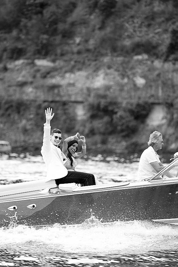 Wedding boat exit with newlyweds on boat waving from a speedboat, groom in white jacket and bride in dress on a lake with trees