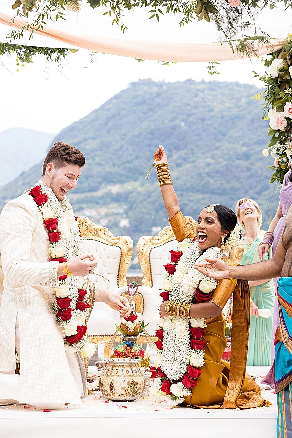 Ceremony moment at an Indian wedding ceremony as bride and groom laugh with red and white garlands beneath a floral canopy on a mountain terrace
