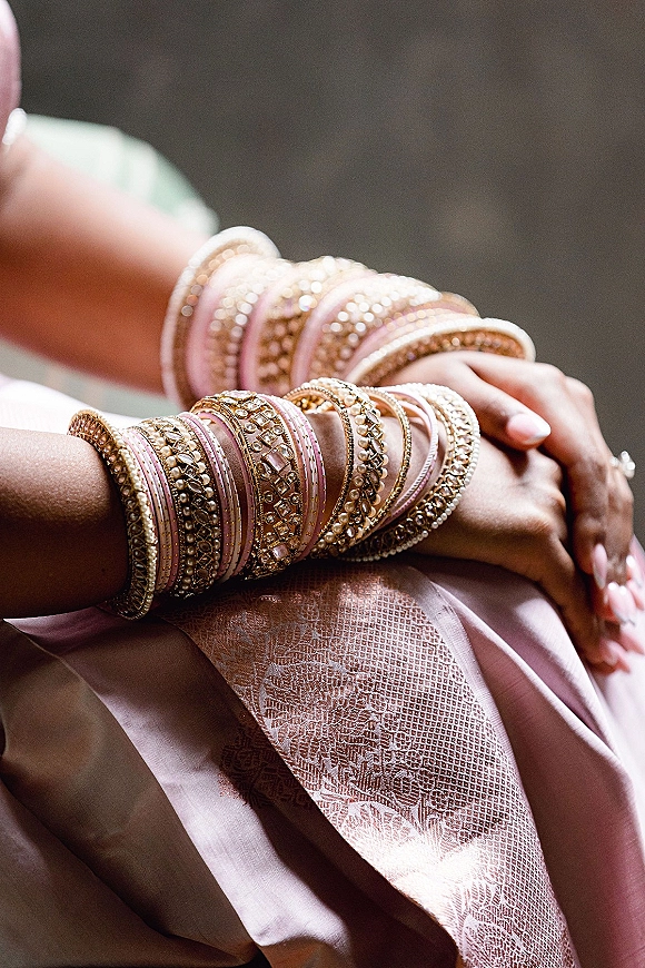 Bridal bangles and indian bridal bangles stacked in pink and gold with pearl accents, wedding ring and manicure against soft bokeh wall