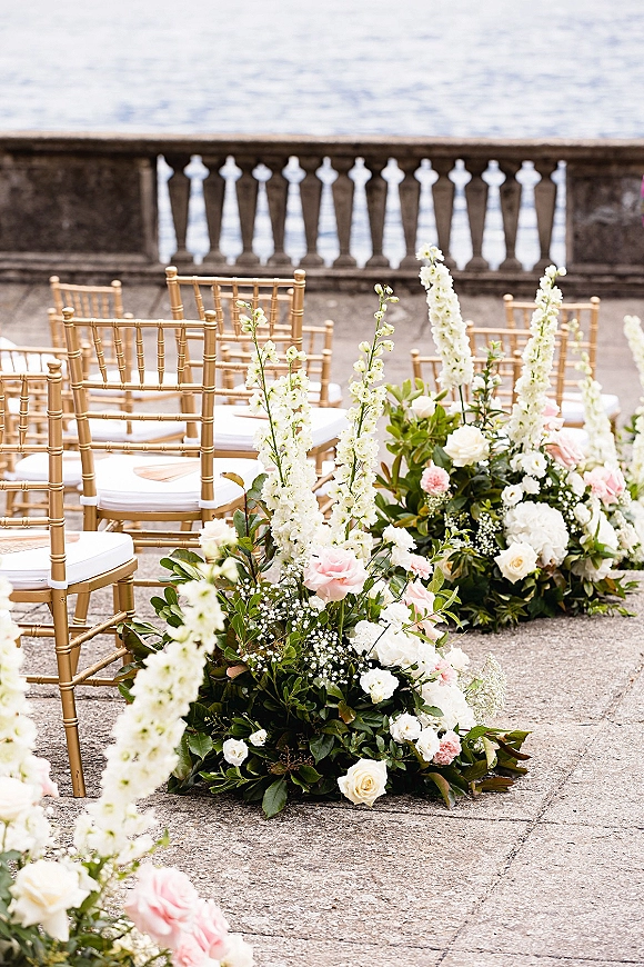 Ceremony aisle decor with outdoor ceremony chairs in gold Chiavari rows, lined with low white and blush roses on an oceanfront stone terrace