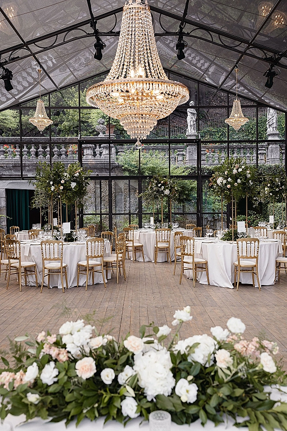 Reception tablescape in a glass tent wedding reception with crystal chandeliers, gold Chiavari chairs, and tall greenery centerpieces in a conservatory setting