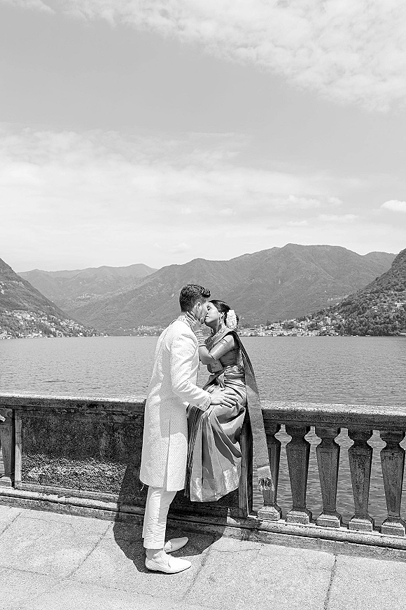 Wedding kiss portrait of a South Asian couple kissing, bride in sari with floral hair garland and groom in sherwani by a mountain lake balustrade