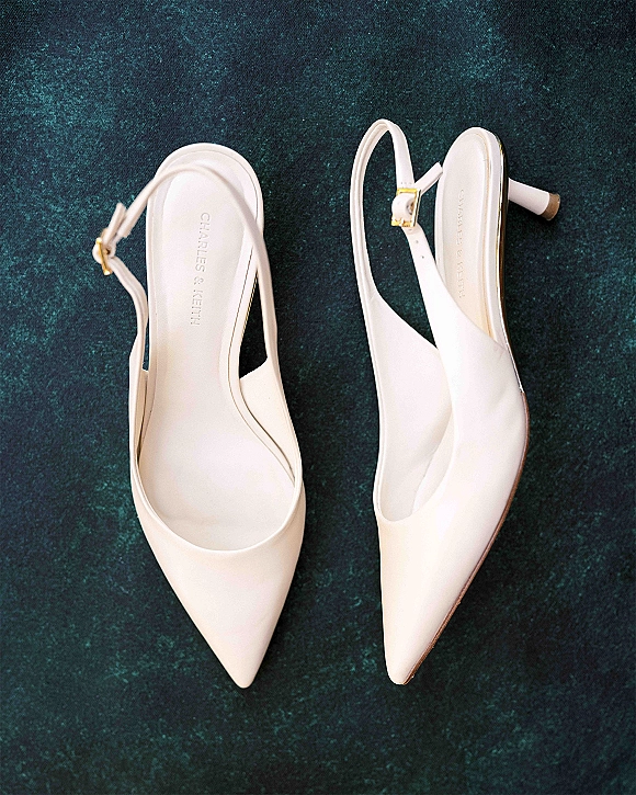 Bridal shoes, white slingback heels with pointed toe, ankle strap and gold buckle, styled on textured dark teal fabric flat lay