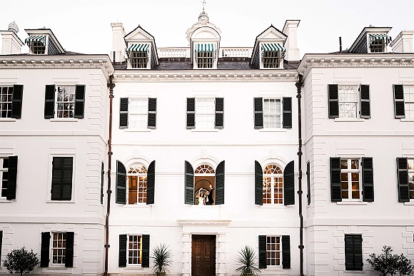 Wedding couple portrait of bride in a strapless dress and groom in a tuxedo waving from an arched window of a white mansion facade
