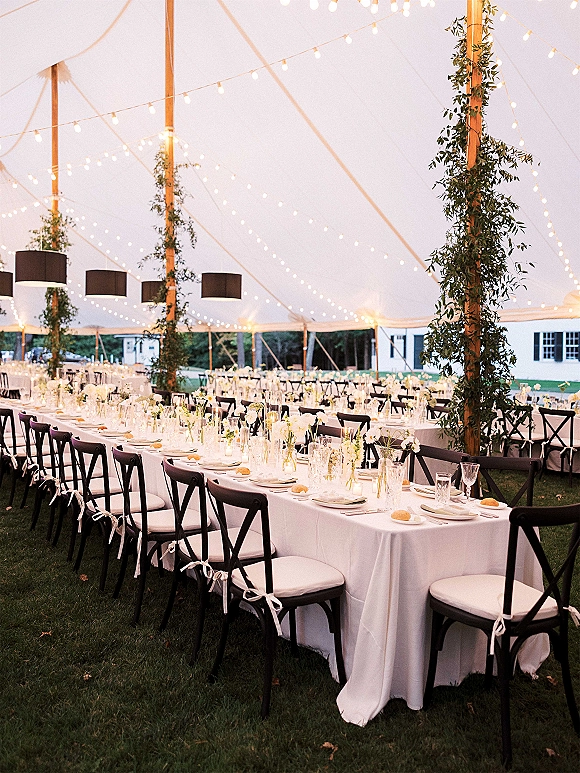 Wedding reception tent with long banquet tables and string lights overhead, set with white linens, candles, and black crossback chairs on a lawn