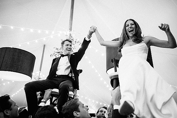 Wedding reception moment as bride and groom lifted on chairs during the hora, holding hands under a tent canopy with string lights overhead