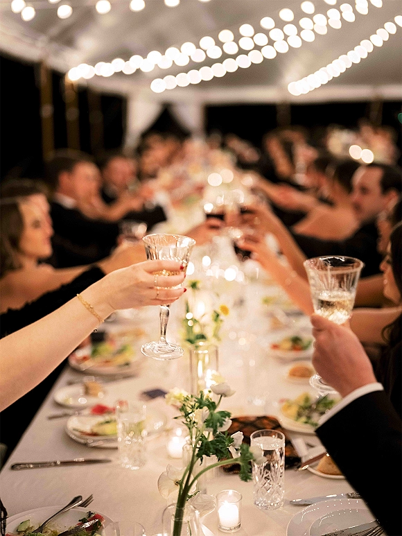 Wedding reception toast as guests cheer with champagne coupes raised at a long table under string lights in a reception tent