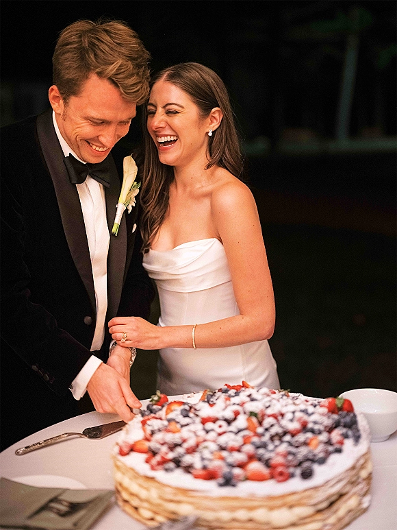 Wedding cake cutting as bride and groom slice a berry-topped cake with powdered sugar, her strapless dress and his tuxedo against a dark reception backdrop