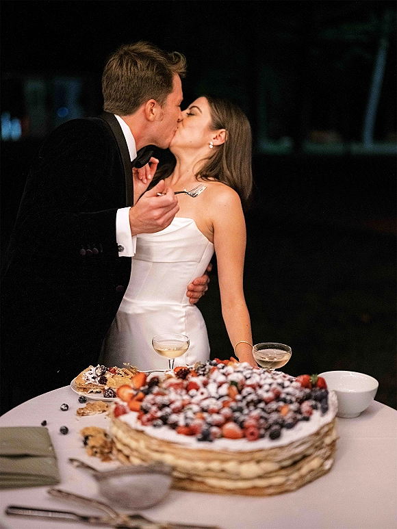 Wedding kiss as bride in strapless dress and groom in tuxedo lean over berry-topped cake at night dessert table with bokeh lights