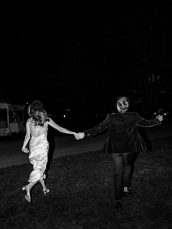 Wedding exit as bride and groom run holding hands across a lawn under string lights by a tent, her heels and dress trailing in the night