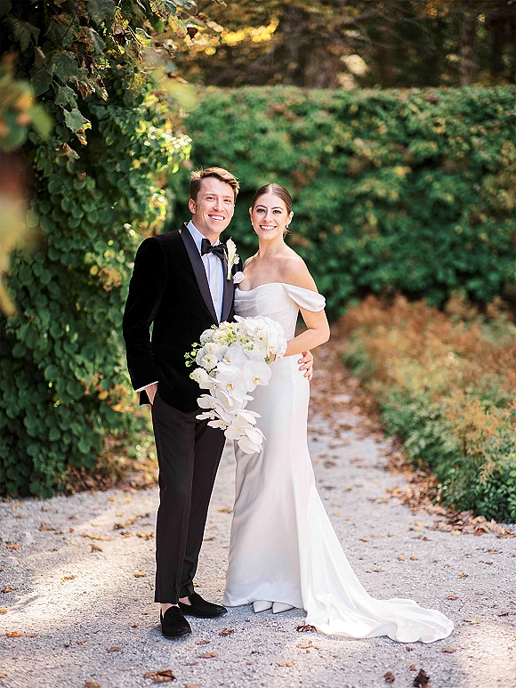 Couple portrait of bride in a strapless gown holding a cascading orchid bouquet beside groom in black tux on a leafy garden path